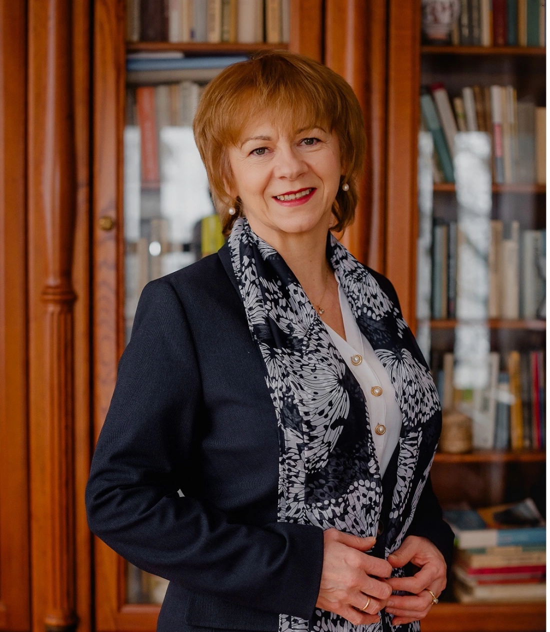 Kobieta w eleganckim ubraniu stoi przed regałem z książkami, uśmiechając się.