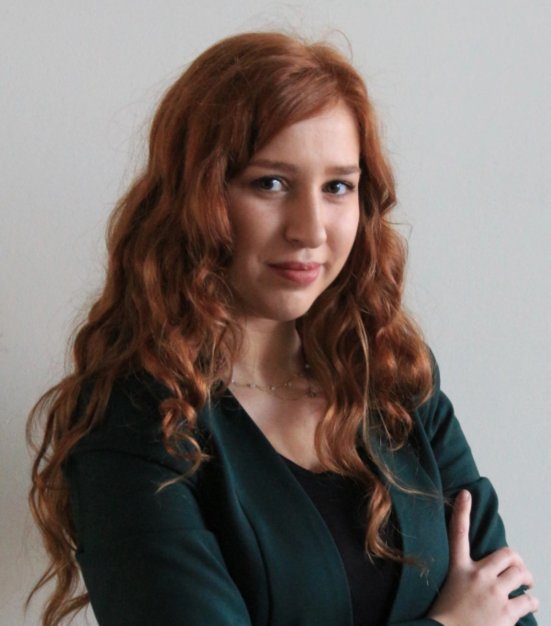 Młoda kobieta z długimi, falowanymi rudymi włosami, ubrana w zielony żakiet, stoi z pewnym wyrazem twarzy.
