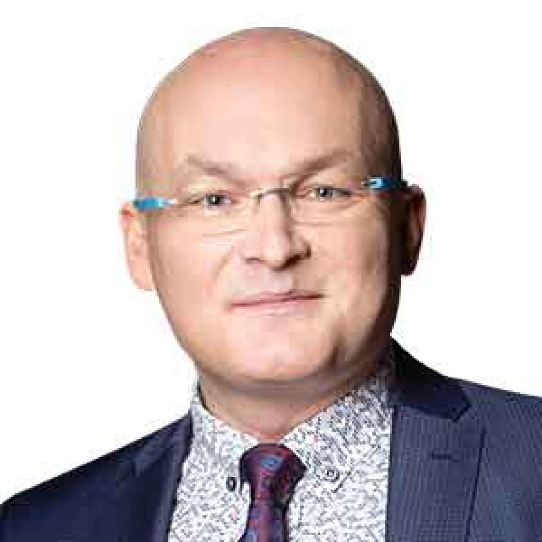 Mężczyzna w okularach z krótkimi włosami, ubrany w garnitur i koszulę w kwiaty.