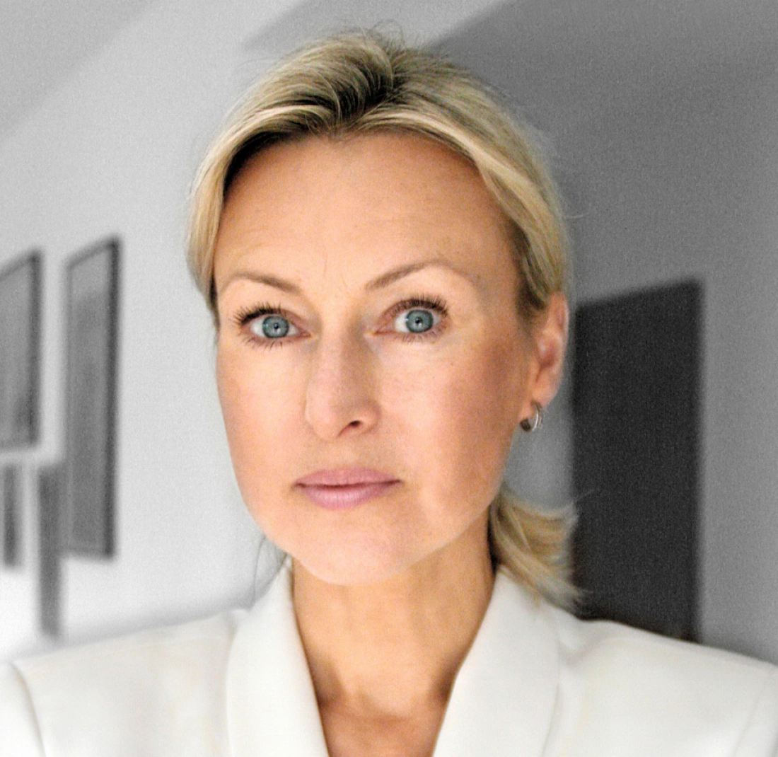 Portret kobiety z blond włosami, o niebieskich oczach, w jasnym ubraniu. W tle delikatnie widoczne kontury wnętrza.