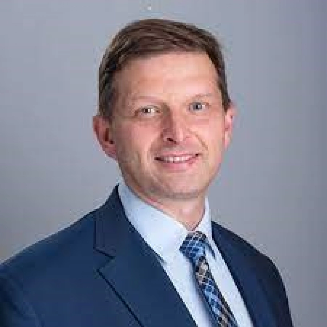 Mężczyzna w garniturze z uśmiechem na tle szarej ściany.