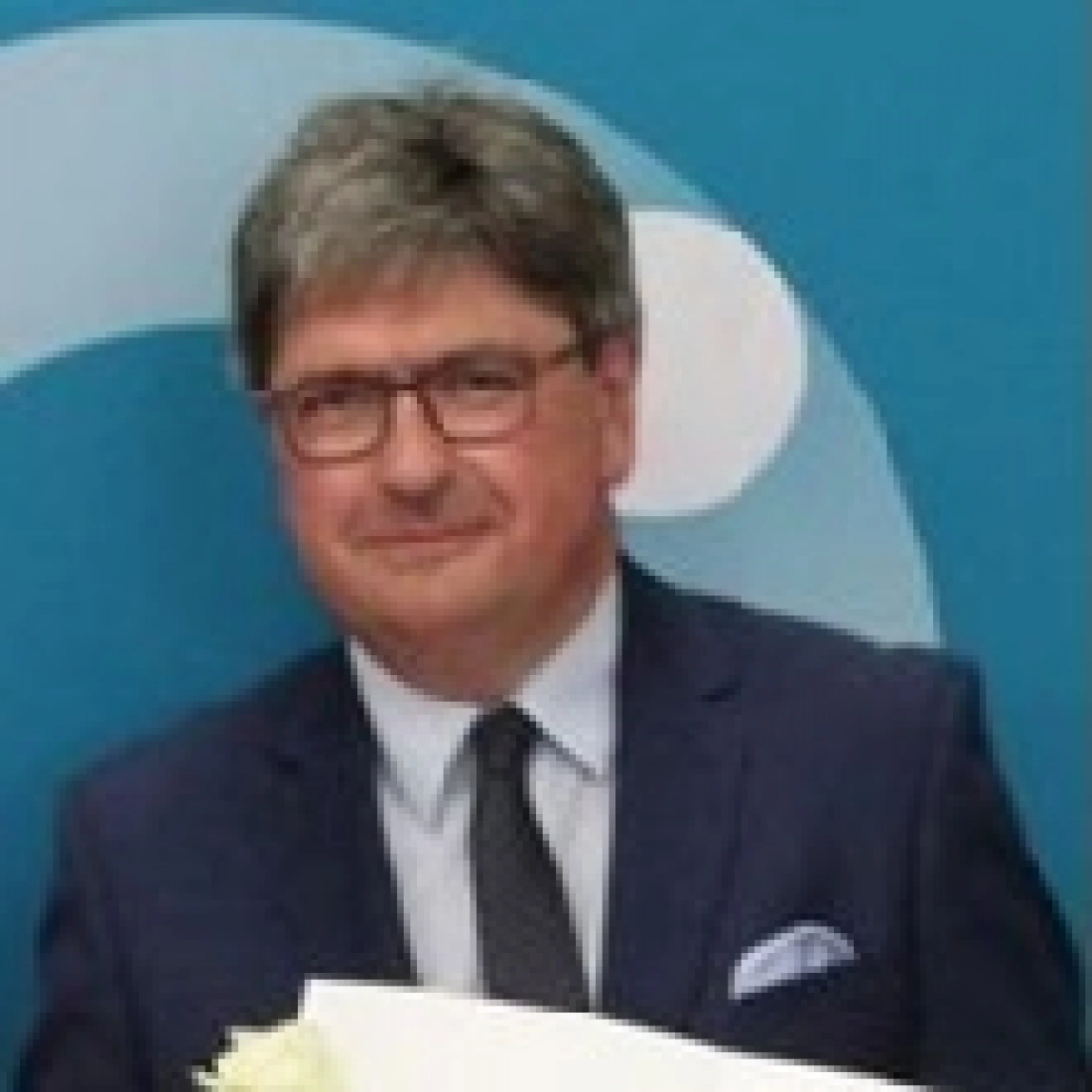 Mężczyzna w garniturze, trzymający dokumenty, na tle niebieskiej ściany.