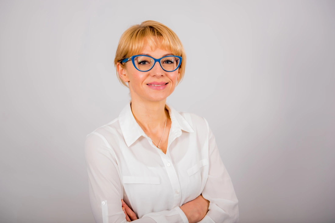 Kobieta w białej koszuli z niebieskimi okularami, uśmiechająca się z założonymi rękami.