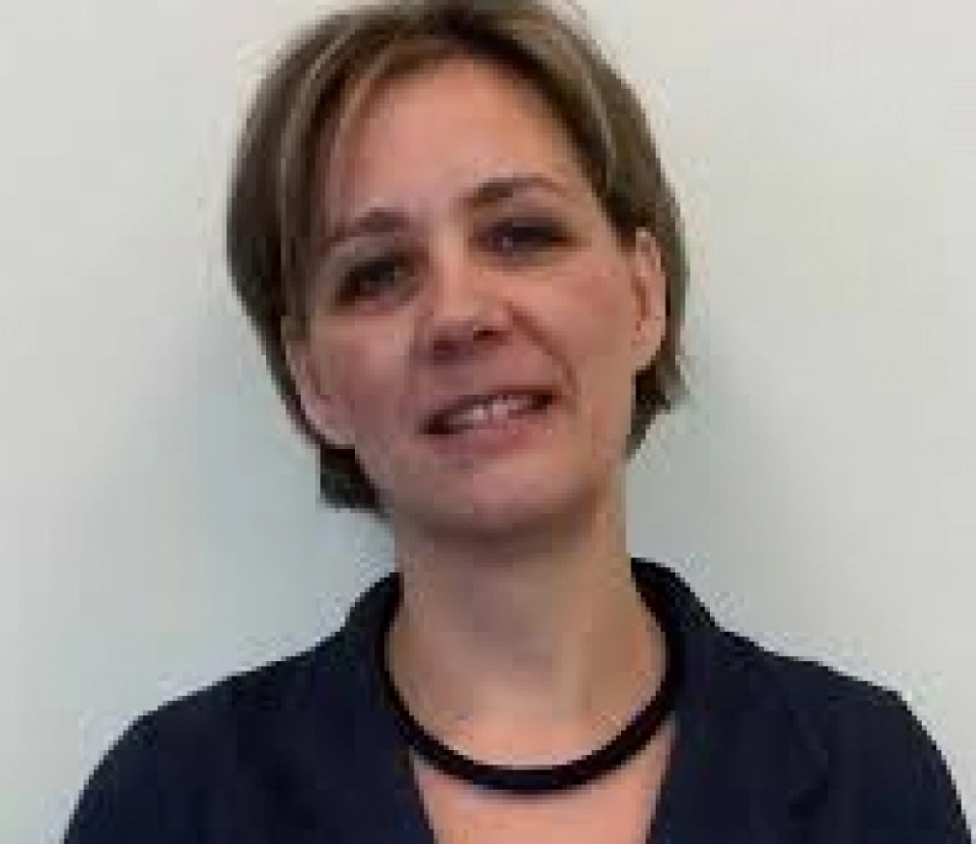 Kobieta w ciemnej bluzce, uśmiechająca się na tle jasnej ściany.