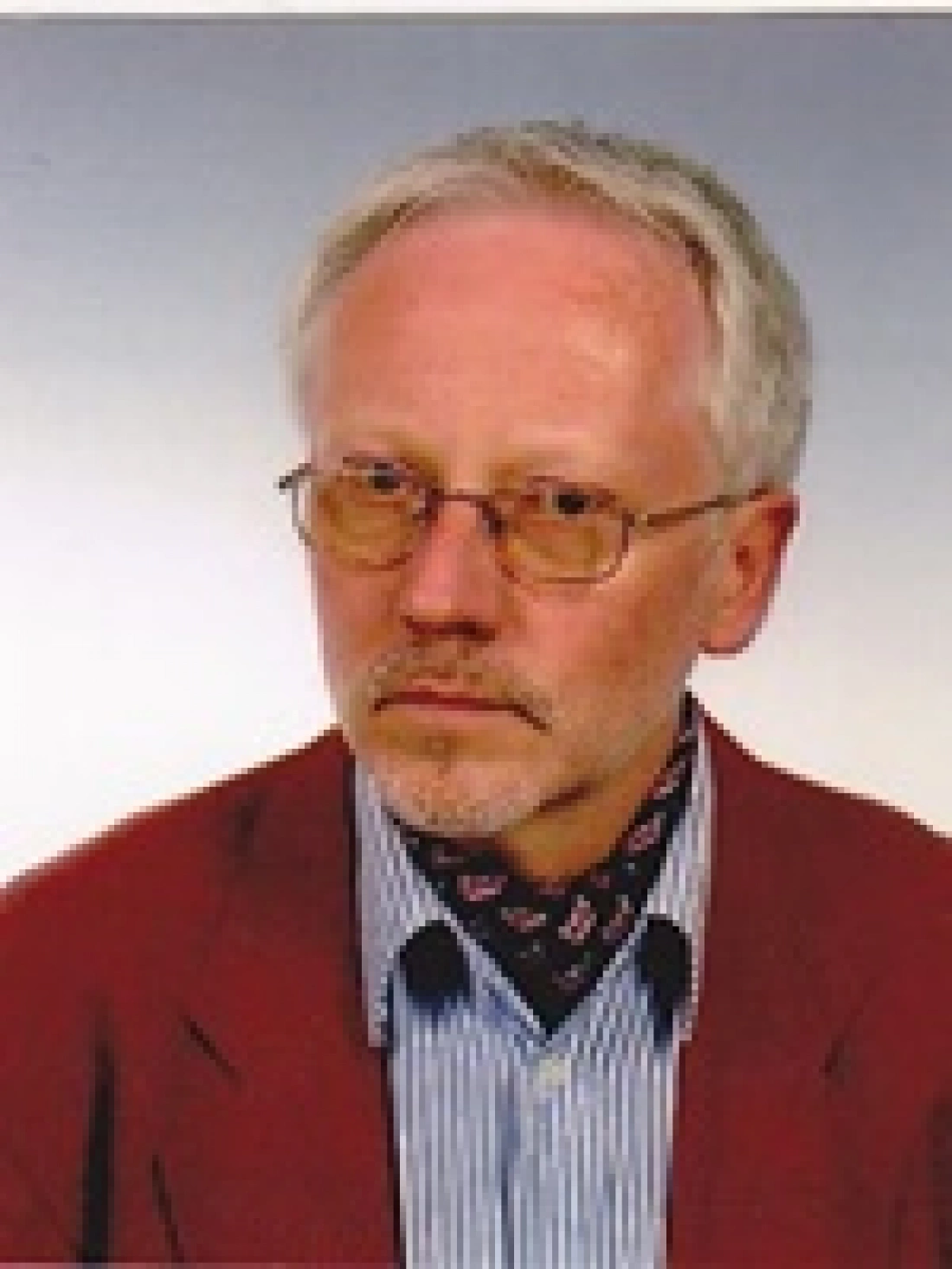 Mężczyzna w okularach, z siwymi włosami i krawatem, ubrany w czerwony żakiet i pasiastą koszulę.