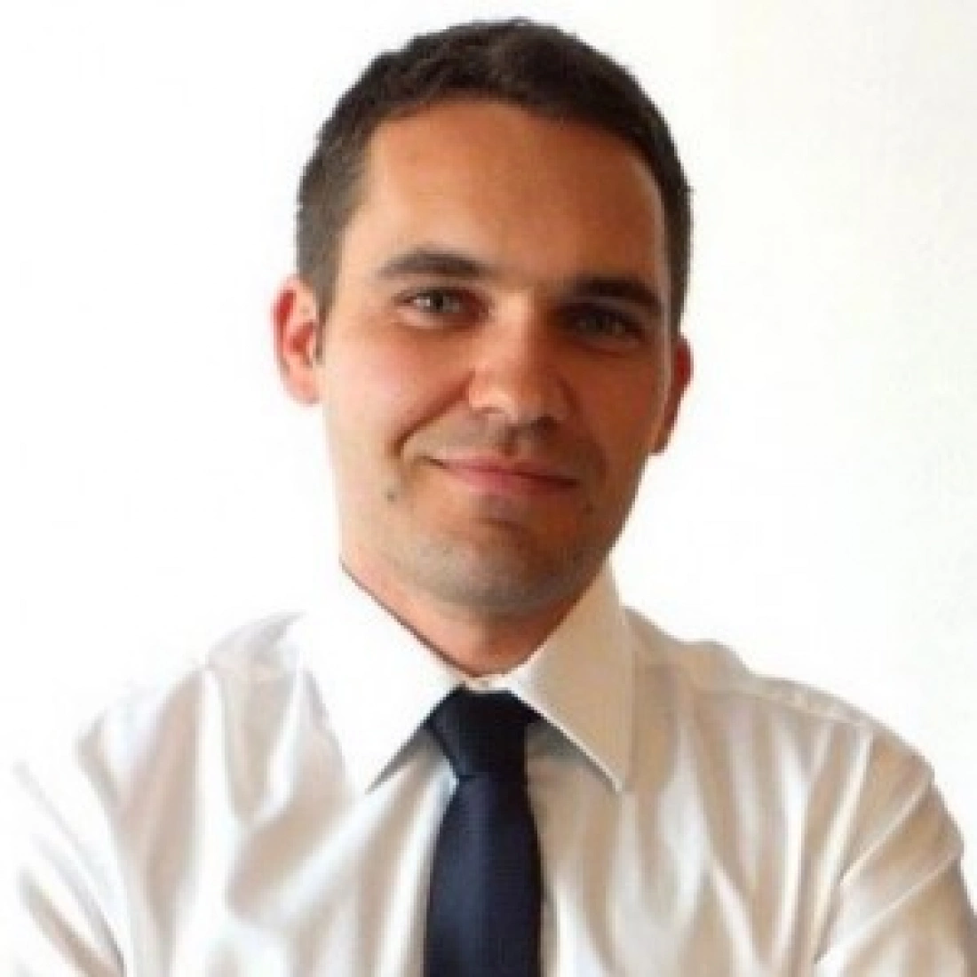 Mężczyzna w białej koszuli i granatowym krawacie, uśmiechający się na tle jasnej ściany.