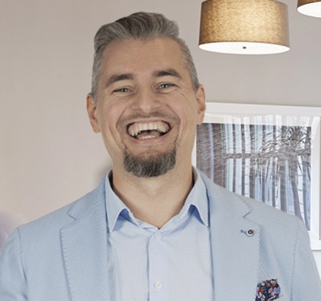 Uśmiechnięty mężczyzna w jasnym garniturze stoi w jasnym pomieszczeniu.