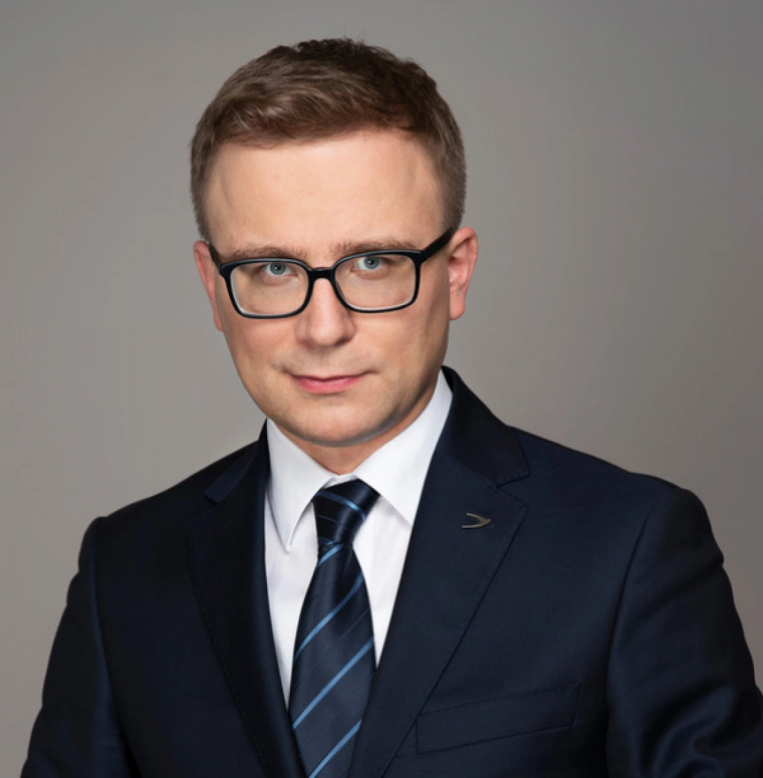 Mężczyzna w okularach, ubrany w elegancki garnitur, pozujący na neutralnym tle.