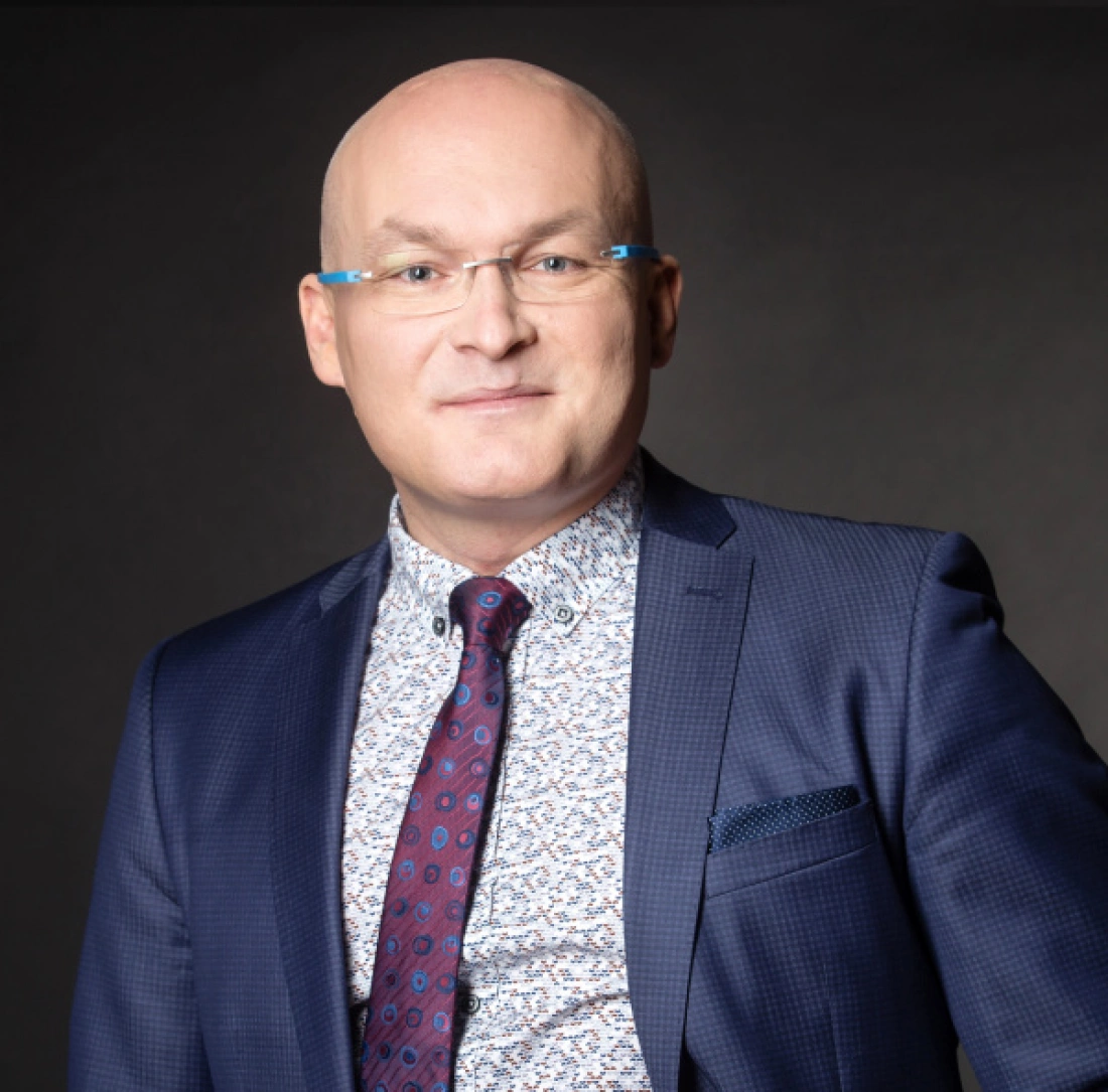 Mężczyzna w okularach, ubrany w garnitur, z uśmiechem na tle ciemnego tła.