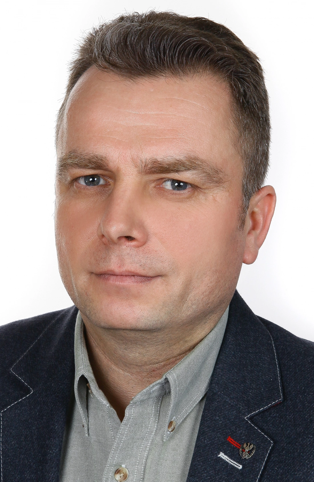 Mężczyzna o krótkich włosach, w szarym, zapinanym na guziki ubraniu oraz ciemnej marynarce, patrzy w kamerę. Tło jest jasne.