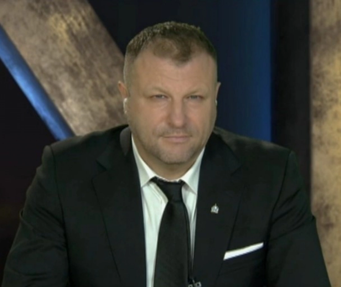 Mężczyzna w garniturze siedzi przy biurku, patrzy prosto w kamerę.