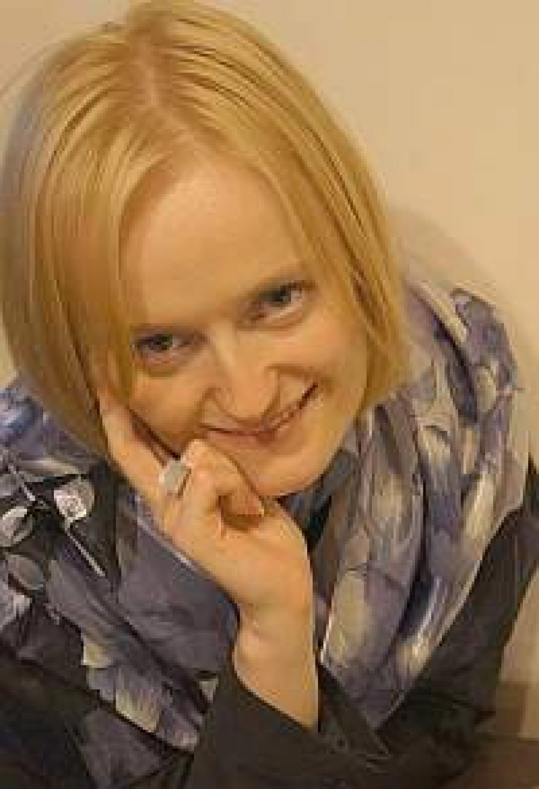 Kobieta z uśmiechem, w niebieskim szaliku, siedząca z ręką przy twarzy.