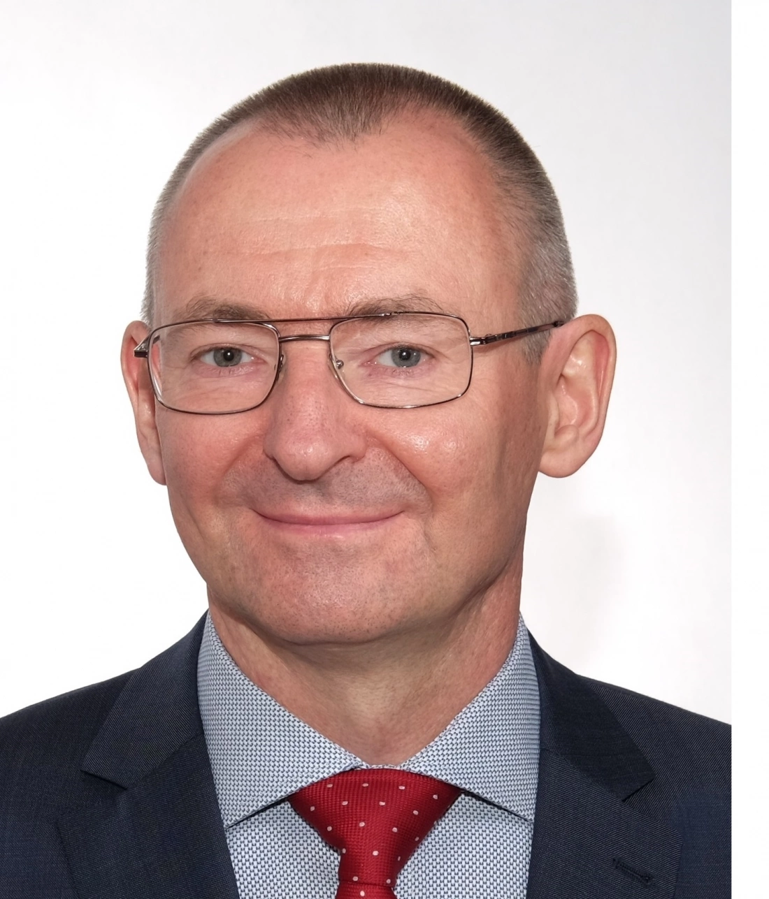 Mężczyzna w garniturze z krawatem, z okularami, uśmiecha się na neutralnym tle.