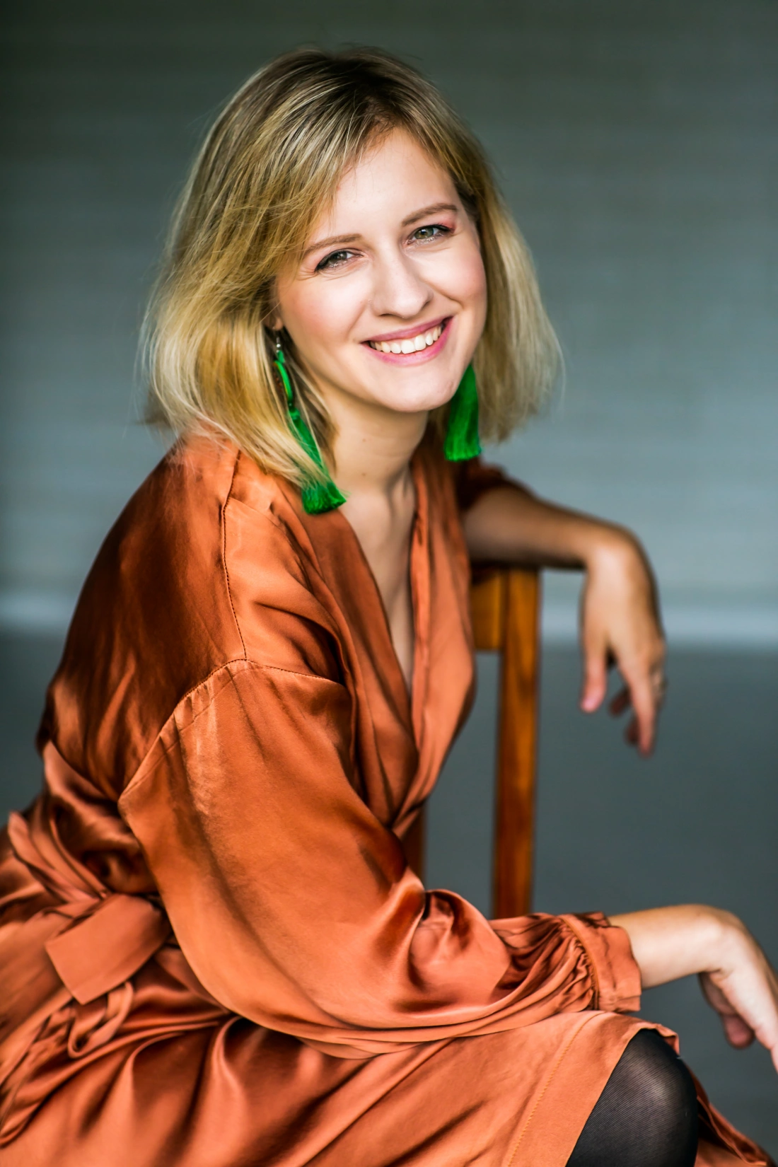 Uśmiechająca się kobieta w pomarańczowej sukience siedzi na krześle, z zielonymi kolczykami.