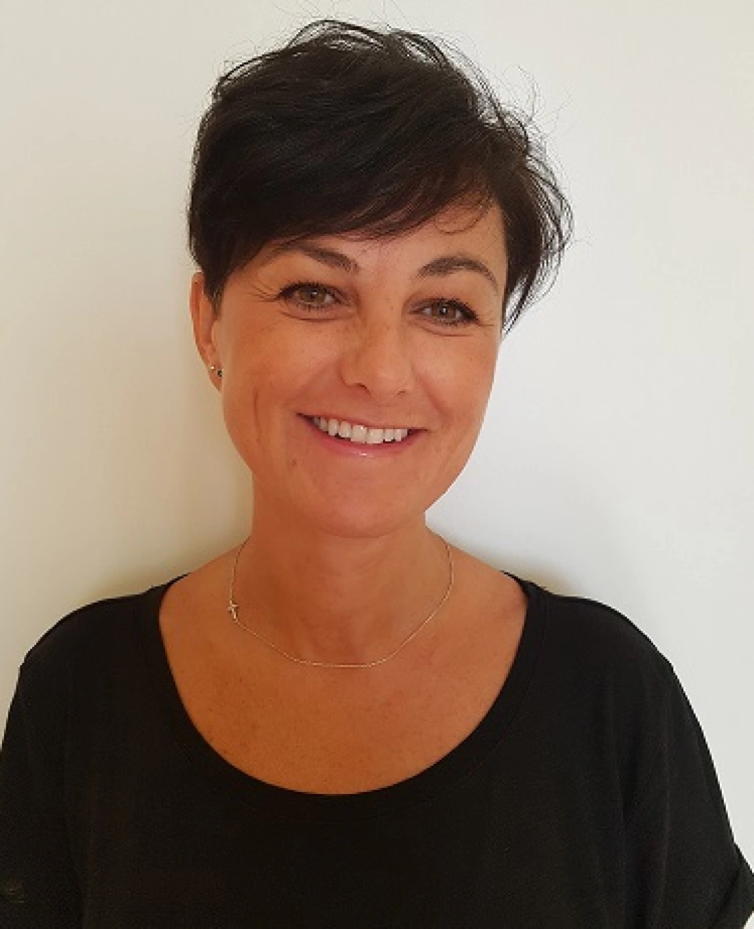 Uśmiechnięta kobieta z krótkimi włosami, ubrana w czarną bluzkę, na tle jasnej ściany.