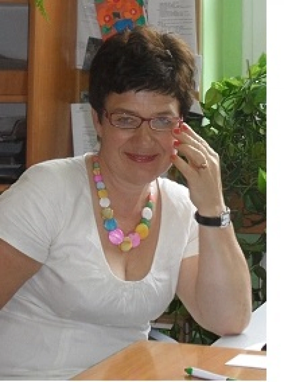 Kobieta w okularach z uśmiechem, siedząca przy biurku, nosi kolorowy naszyjnik.