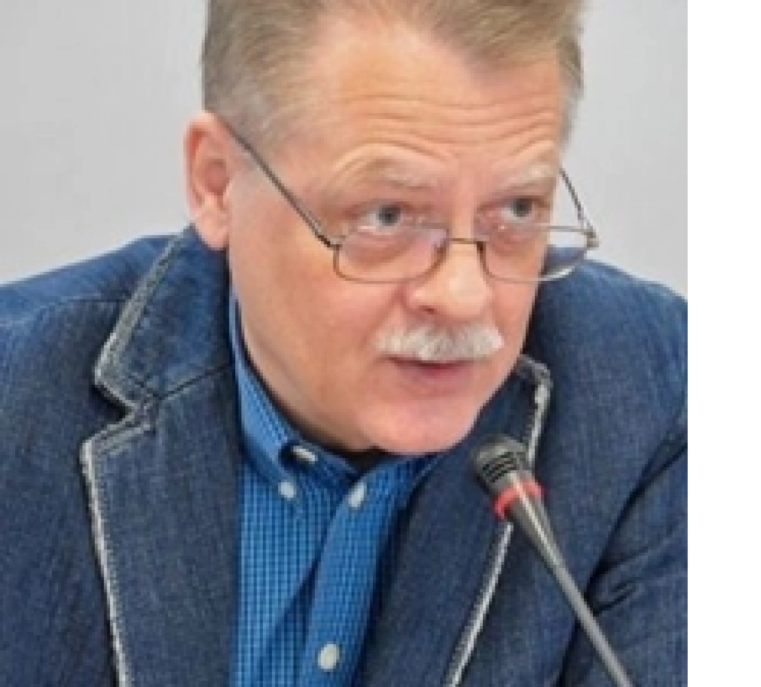 Mężczyzna w okularach, ubrany w niebieską koszulę i dżinsową marynarkę, mówi do mikrofonu.