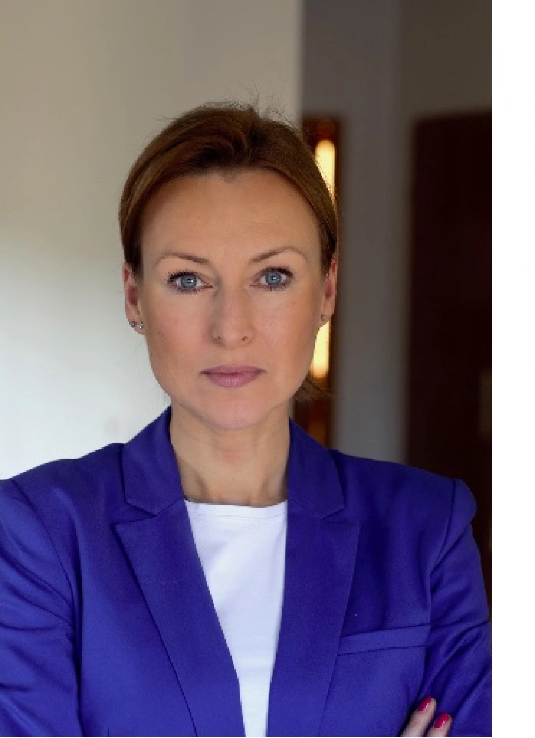 Kobieta w fioletowej marynarce, z poważnym wyrazem twarzy, stoi z założonymi rękami.
