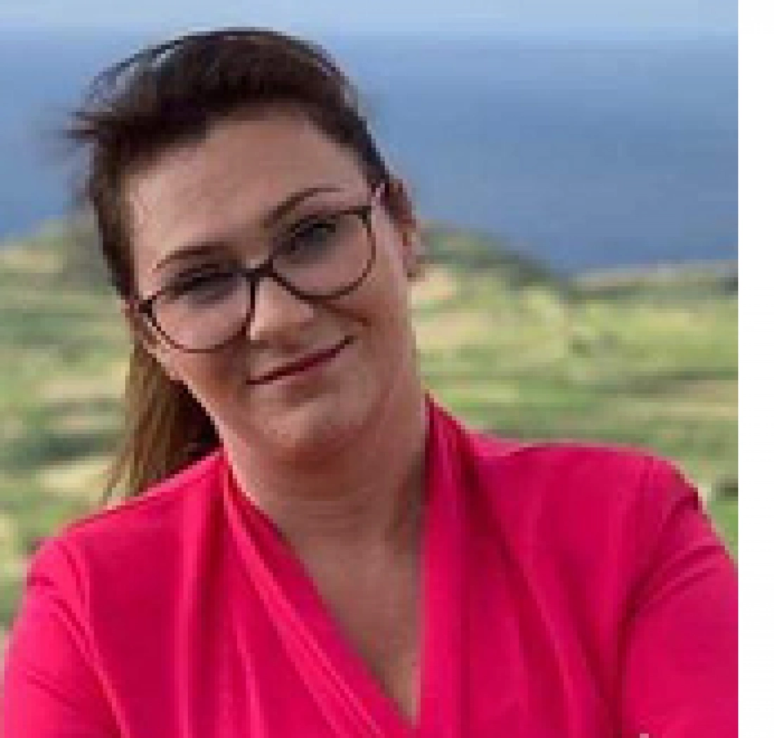 Uśmiechnięta kobieta w różowej bluzce na tle zielonych wzgórz i nieba.