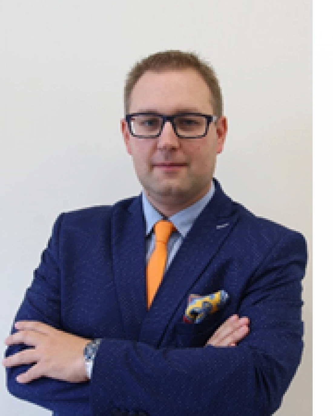 Mężczyzna w garniturze z pomarańczowym krawatem, stojący z założonymi rękami.