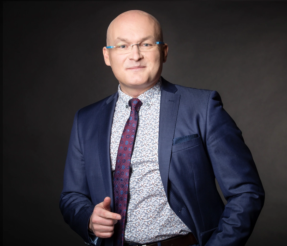 Mężczyzna w garniturze z krawatem, uśmiechający się i wskazujący palcem.