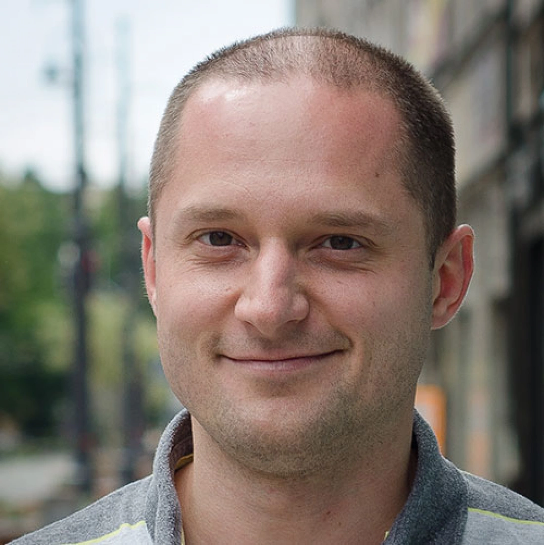 Mężczyzna z krótkimi włosami stoi na tle ulicy, uśmiechając się.