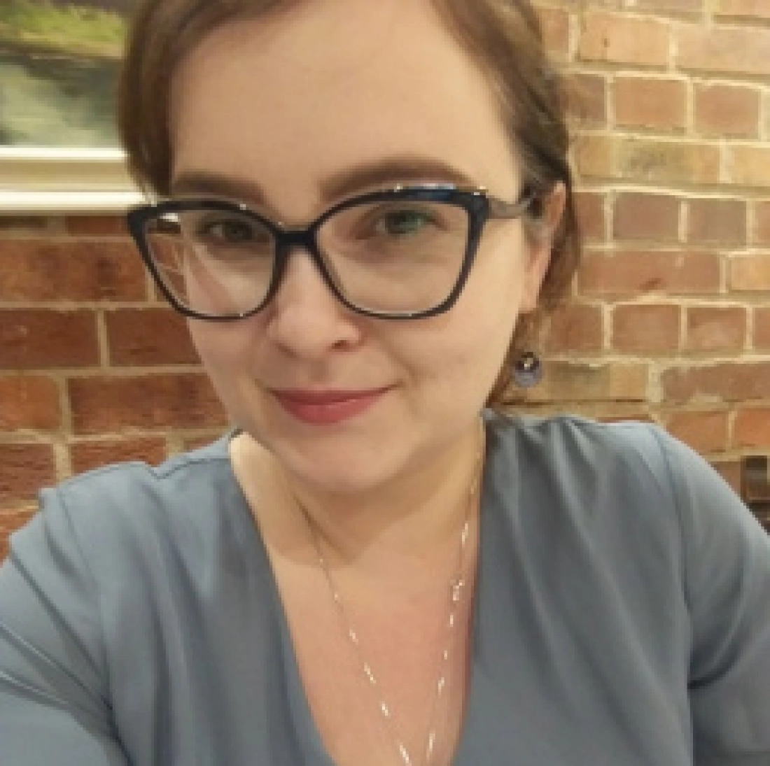 Kobieta z okularami, uśmiechająca się przy ceglanej ścianie.