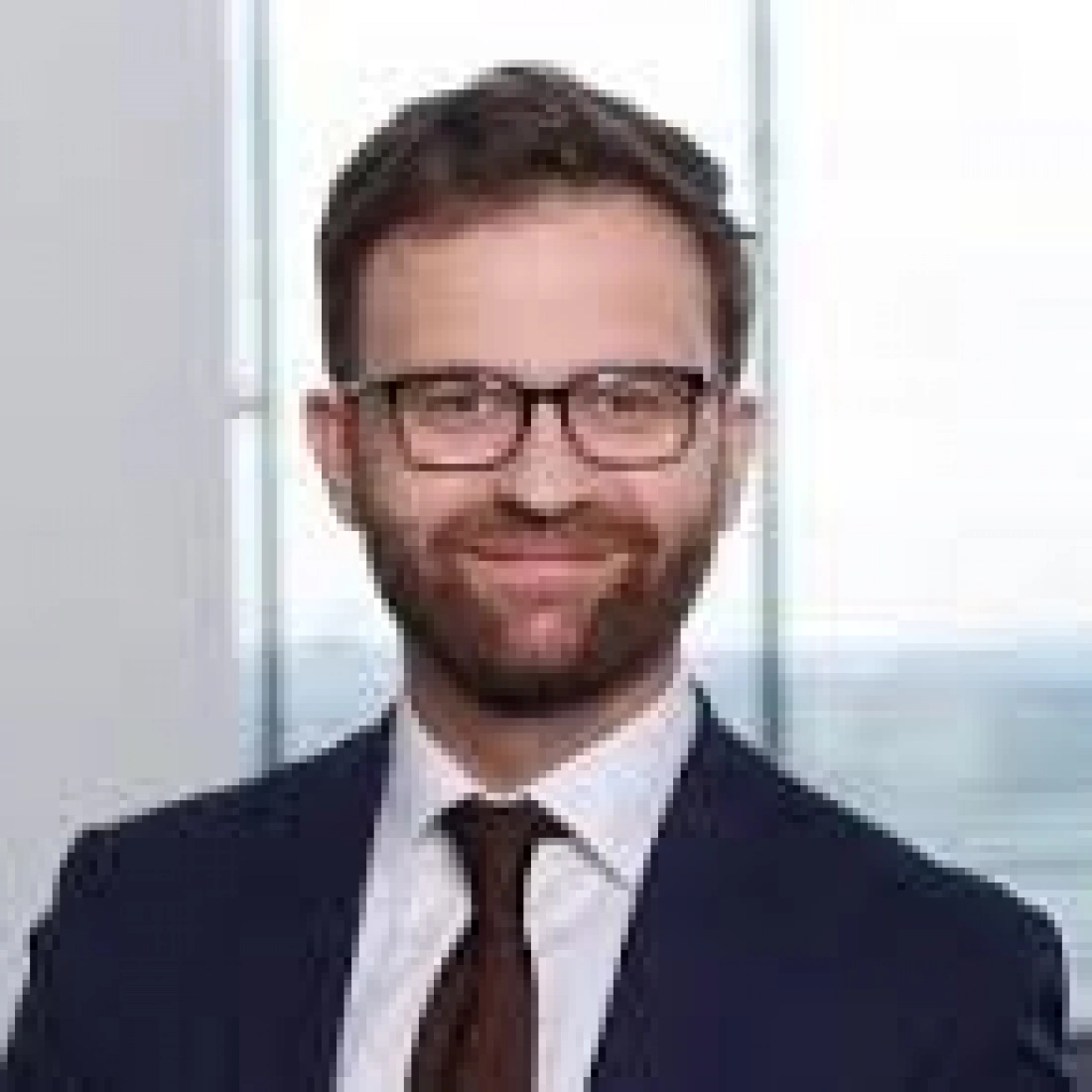 Mężczyzna w okularach, uśmiechający się w garniturze, stojący przy oknie.