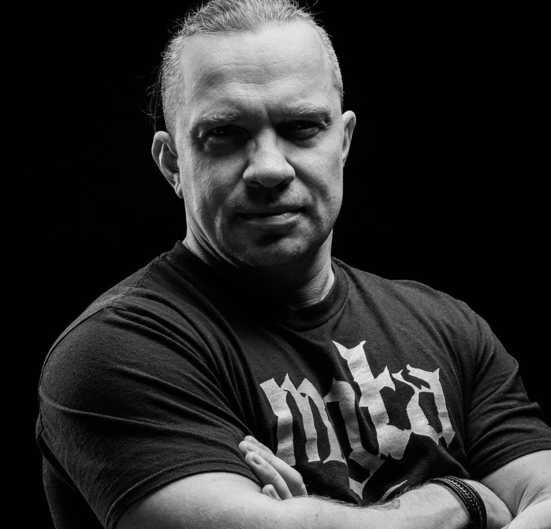 Mężczyzna w ciemnym tle, z założonymi rękami i poważnym wyrazem twarzy, nosi czarny t-shirt z dużym napisem.