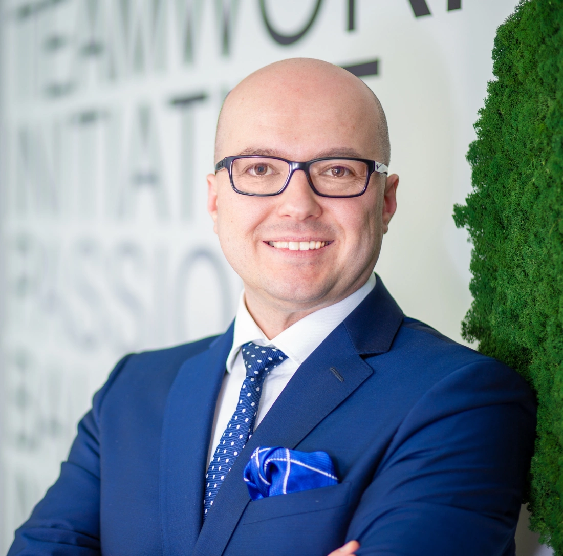 Mężczyzna w okularach, ubrany w niebieski garnitur z krawatem, uśmiecha się, stojąc obok zielonej rośliny. W tle widoczne napisy związane z pracą.