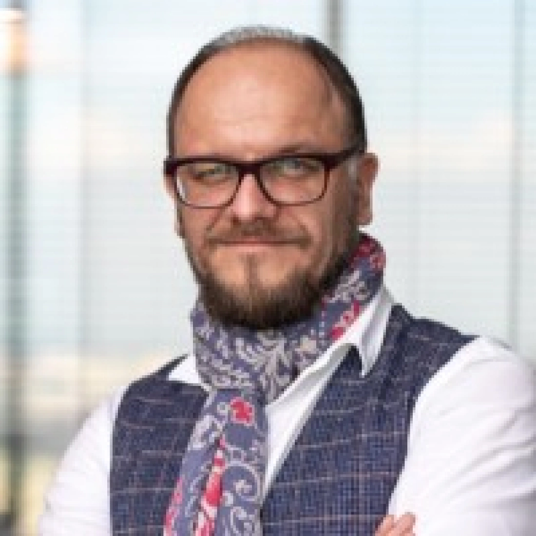 Mężczyzna w okularach, z brodą i apaszką, stoi z założonymi rękami, uśmiechając się. Tło z oknem i widokiem na miasto.