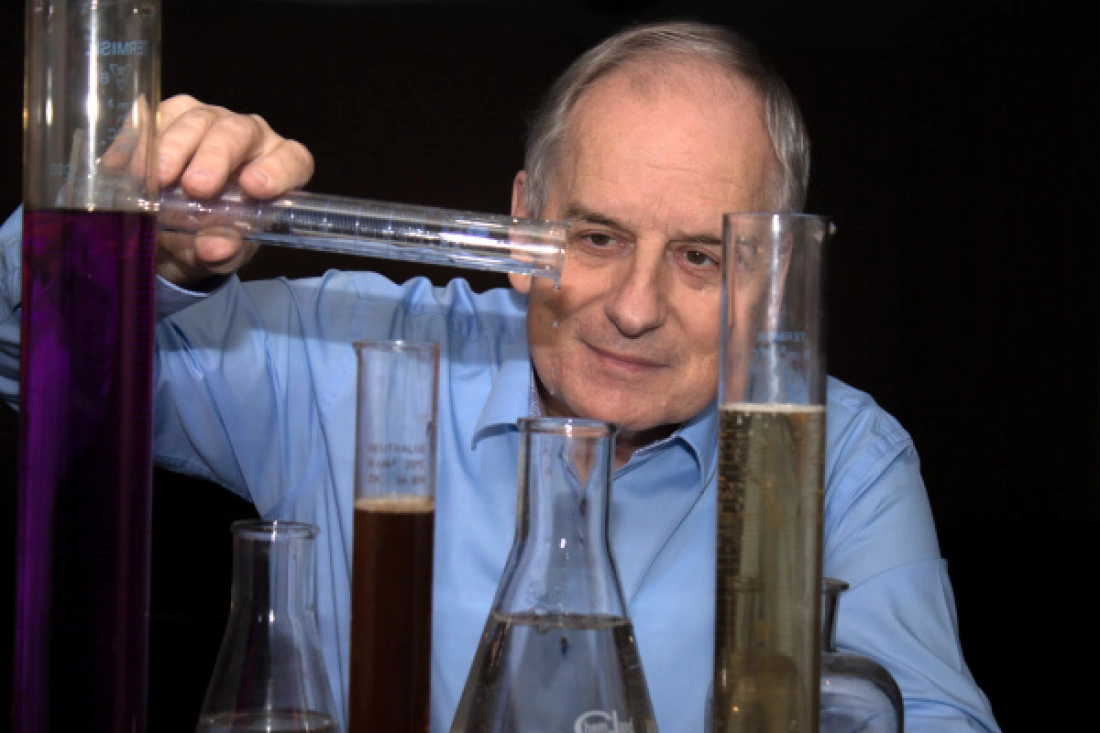 Mężczyzna w niebieskiej koszuli przelewa ciecz z jednego cylindra do drugiego, otoczony różnymi probówkami i naczyniami laboratoryjnymi.