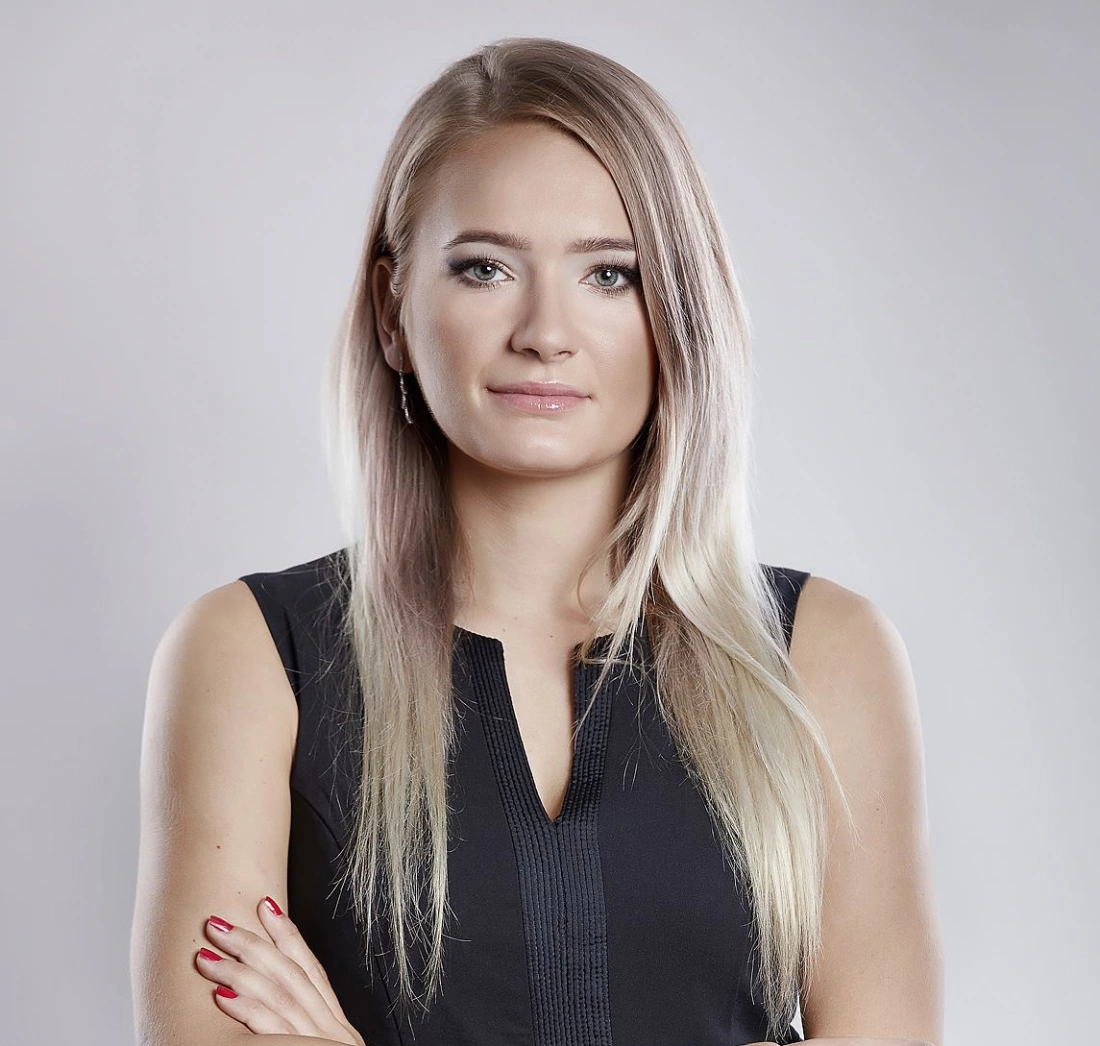 Kobieta z długimi, jasnymi włosami, ubrana w czarną bluzkę, stoi na neutralnym tle z pewnym wyrazem twarzy.