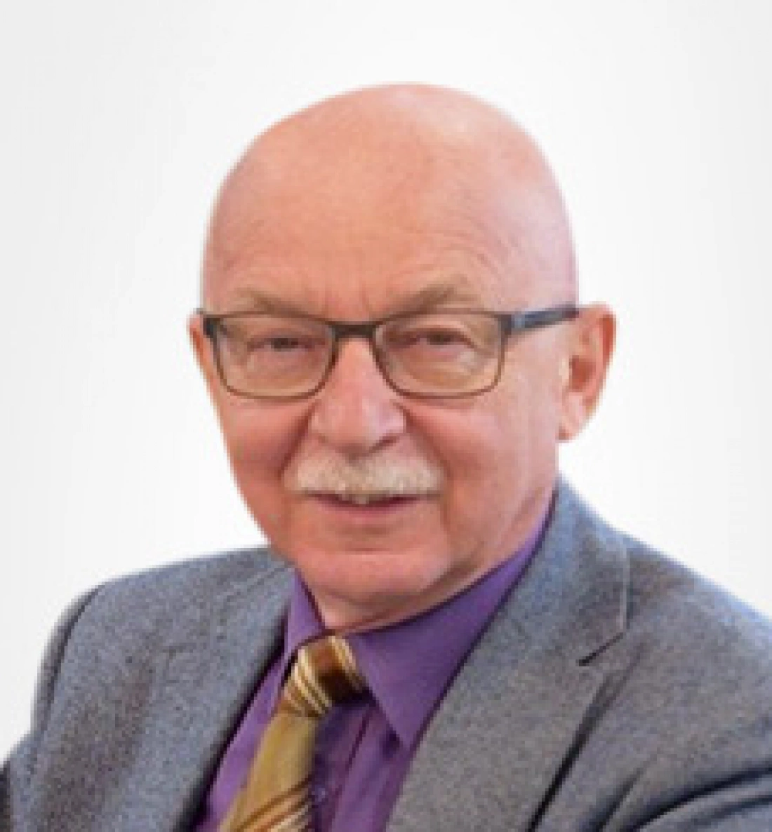 Mężczyzna w okularach z siwą brodą, ubrany w szary garnitur i fioletową koszulę.