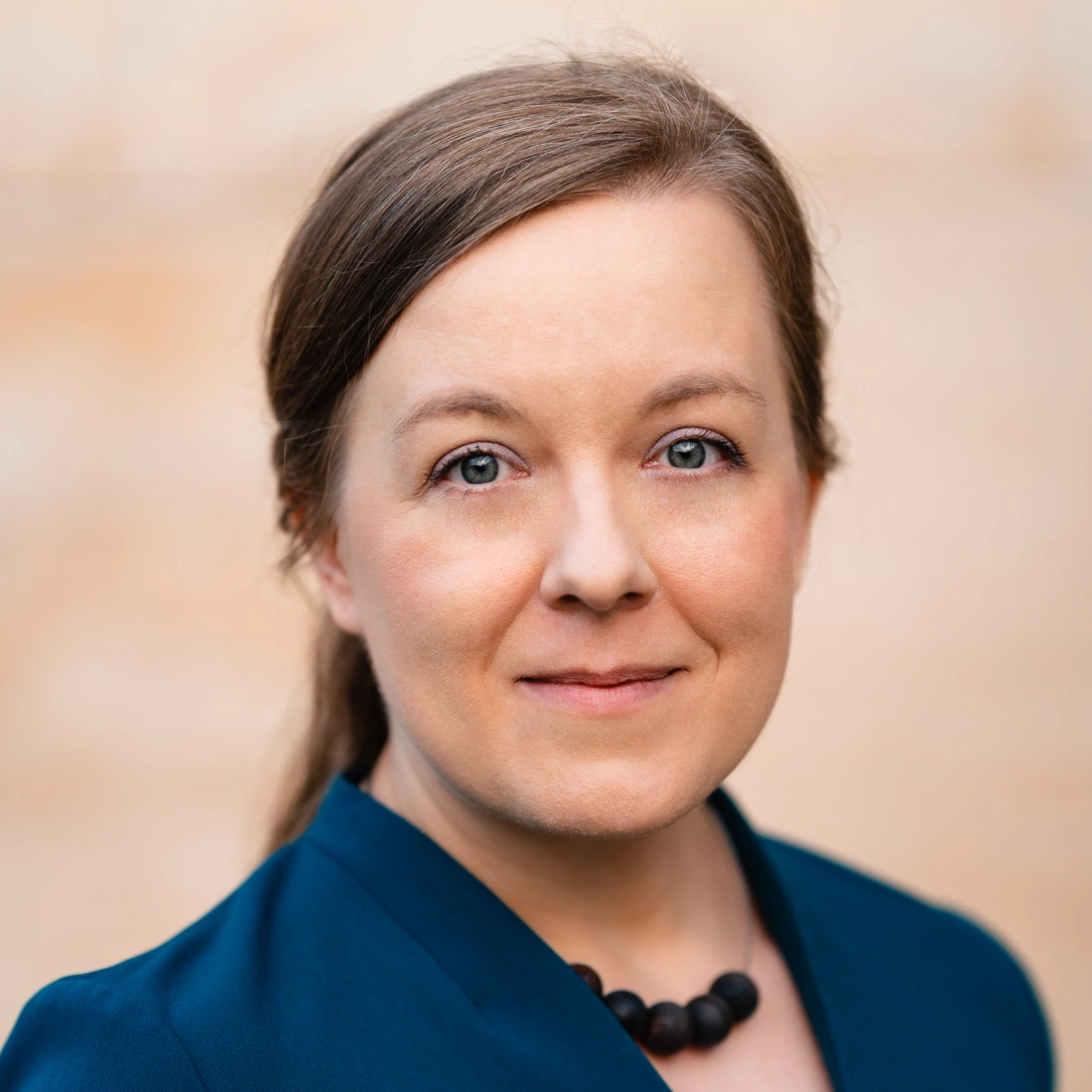 Portret młodej kobiety o jasnych oczach, w granatowym żakiecie i naszyjniku, na tle jasnej ściany.