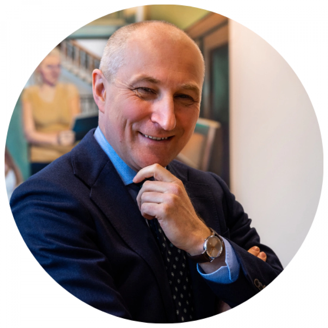 Mężczyzna w garniturze z uśmiechem, stojący w półprofilu, z ręką przy brodzie. W tle widoczne obrazy.