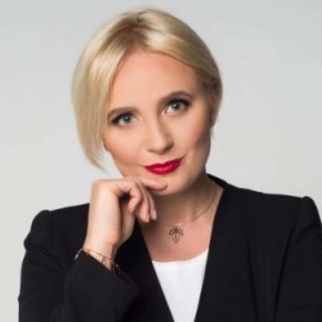 Kobieta w eleganckim żakiecie, z krótkimi blond włosami, uśmiechająca się i trzymająca dłoń przy brodzie.