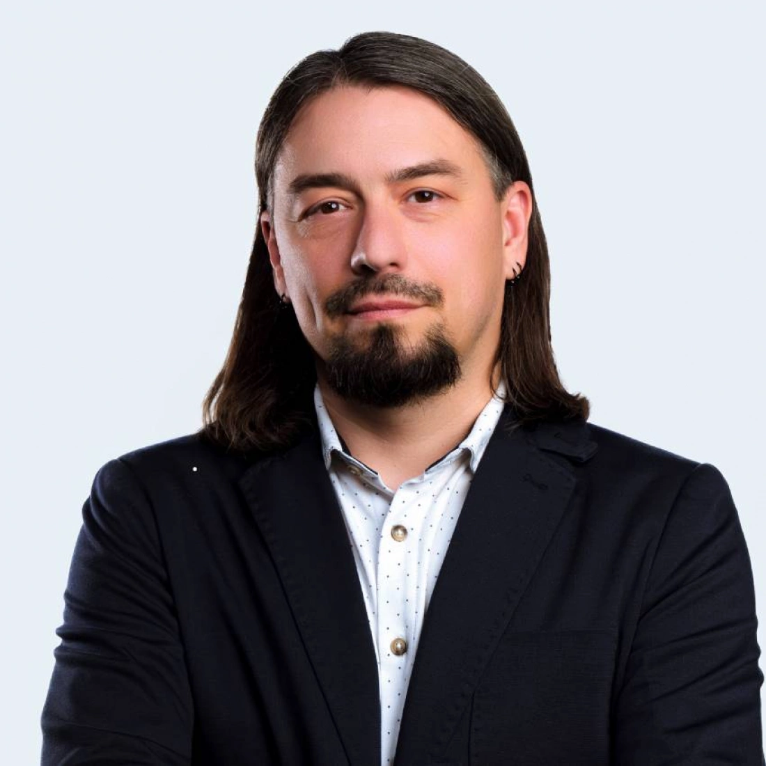 Mężczyzna z długimi włosami i zarostem, ubrany w ciemny garnitur i białą koszulę w kropki, pozujący na jasnym tle.