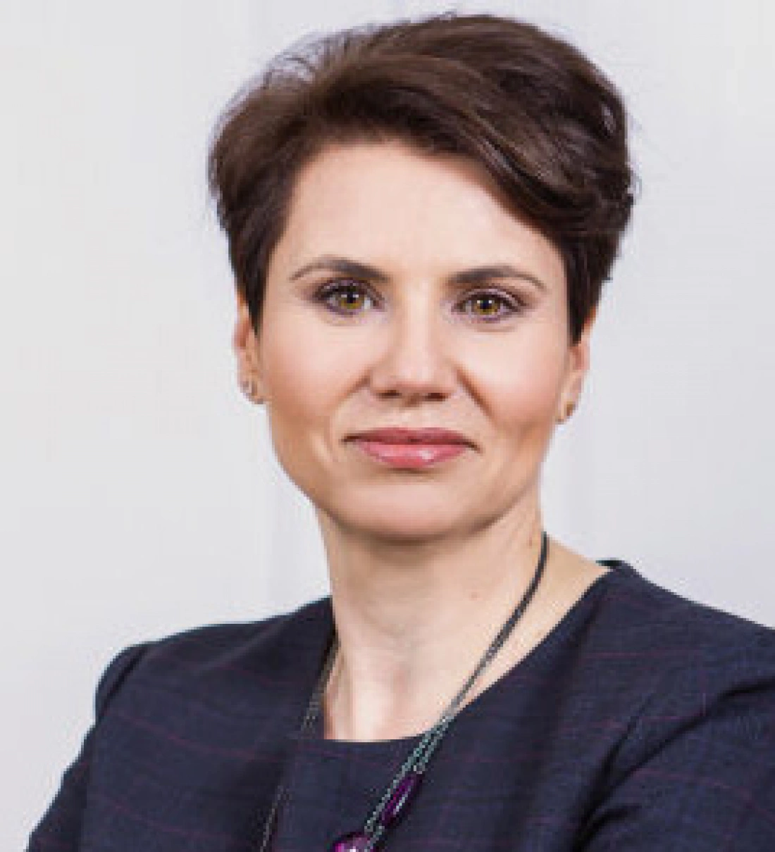 Kobieta o krótkich włosach, ubrana w elegancki strój, patrzy w kamerę z poważnym wyrazem twarzy.