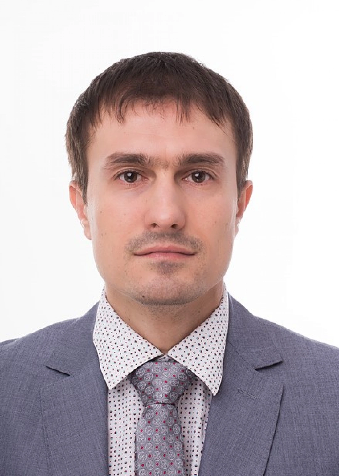 Mężczyzna w garniturze z krawatem, o neutralnym wyrazie twarzy, na jasnym tle.