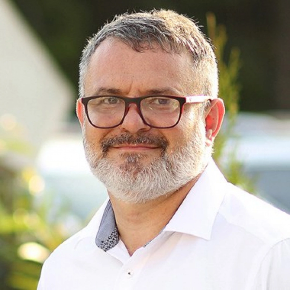 Mężczyzna z zarostem i okularami, uśmiechający się w jasnej koszuli w plenerze.