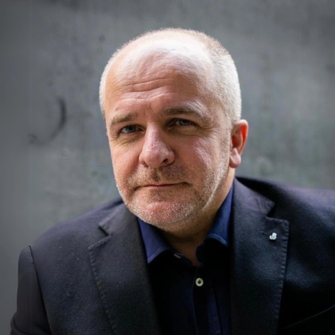 Mężczyzna o krótkich, siwych włosach, ubrany w ciemną marynarkę, stoi na tle szarej ściany.