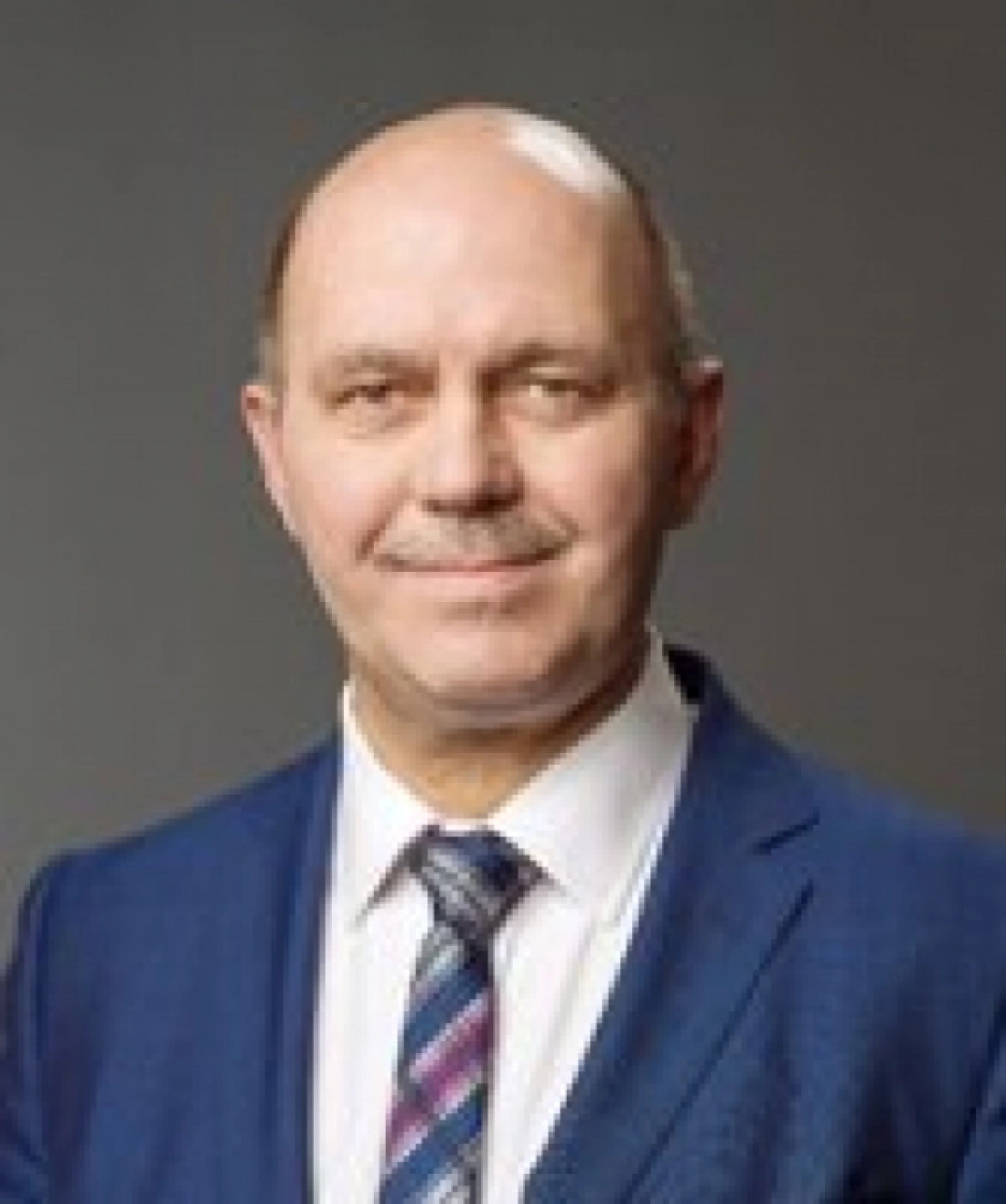 Mężczyzna w garniturze, z uśmiechem, na tle szarej ściany.