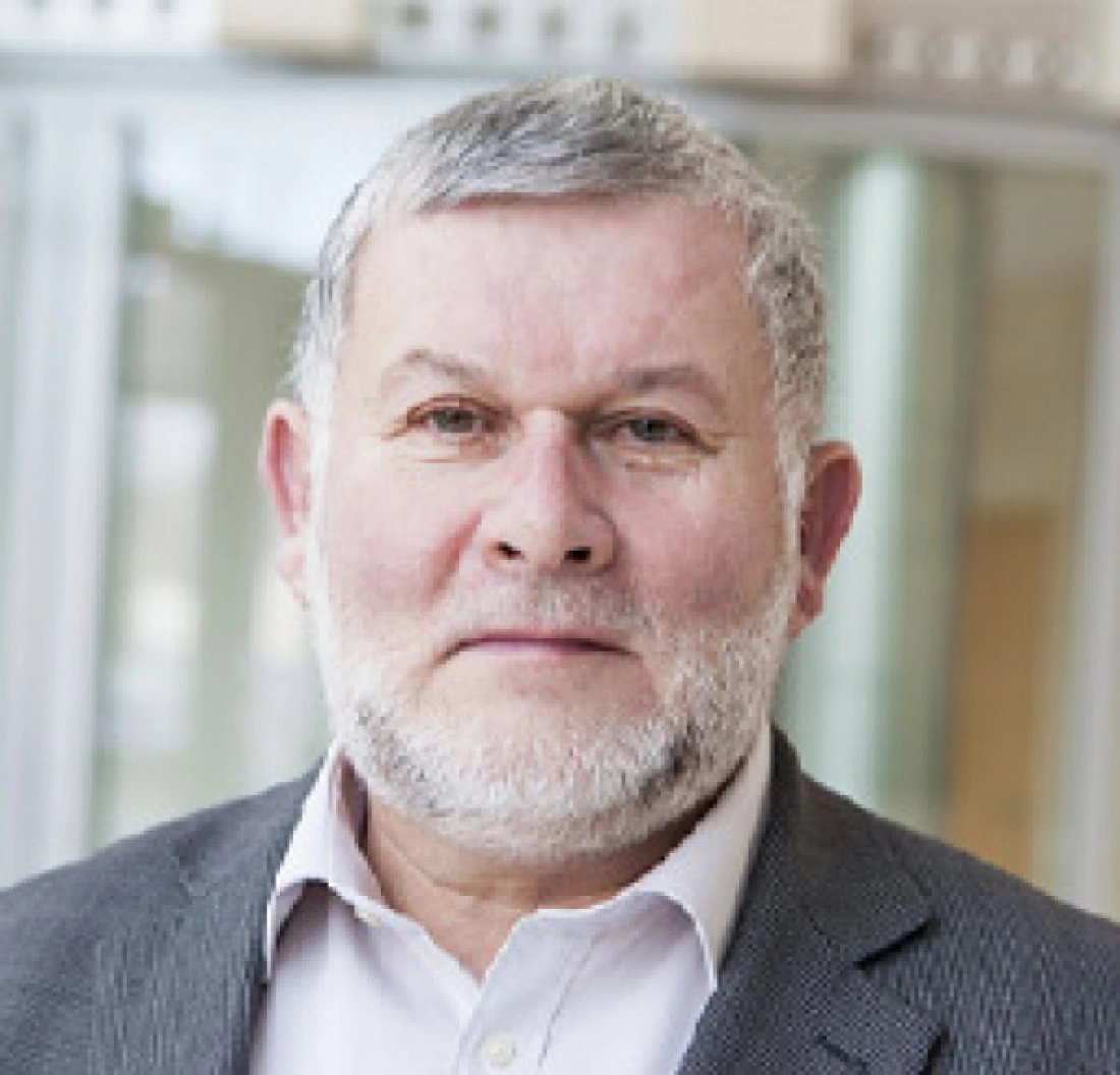 Mężczyzna z siwą brodą, ubrany w szary żakiet, pozujący do zdjęcia w jasnym otoczeniu.