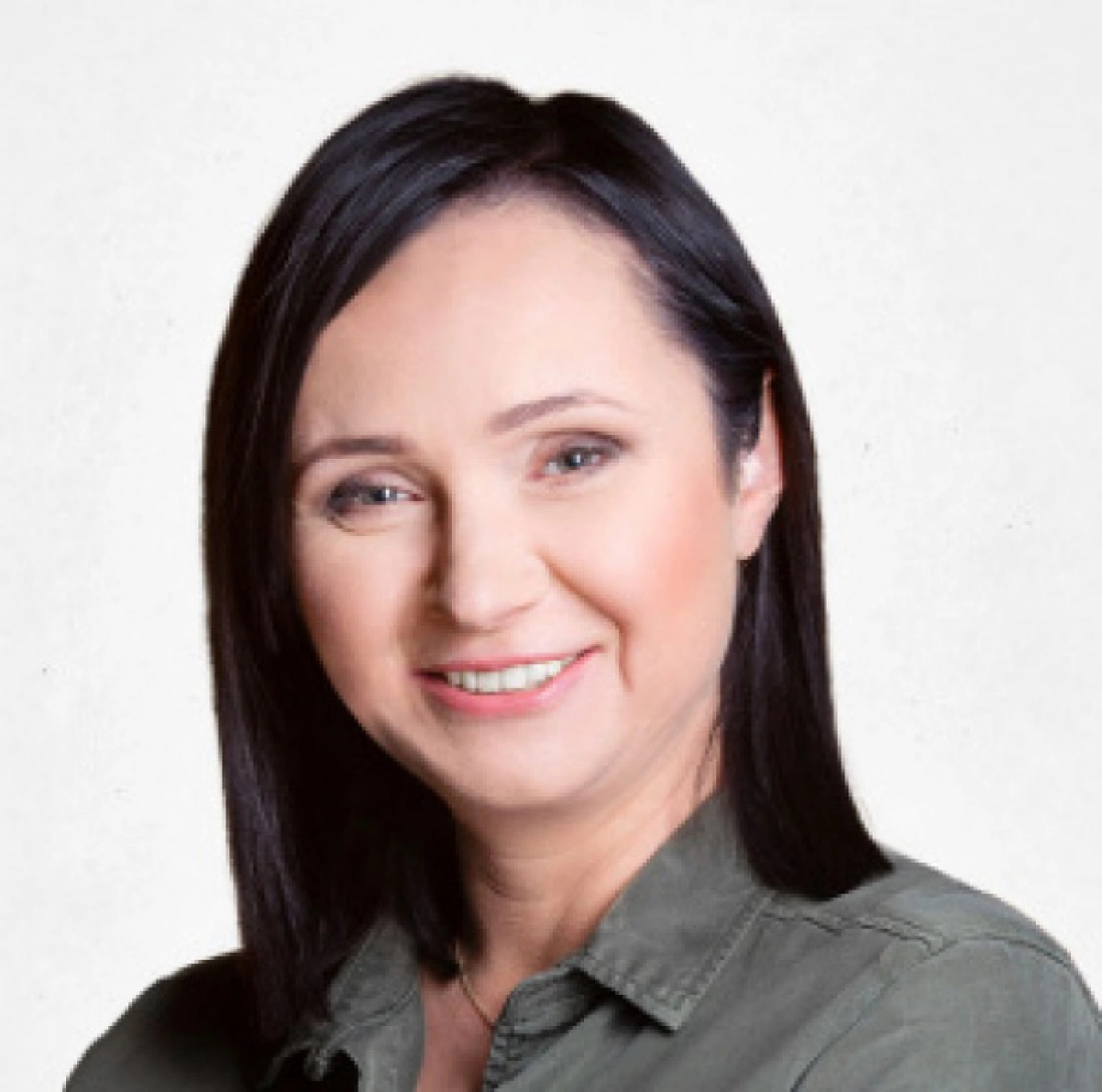 Uśmiechnięta kobieta z długimi, ciemnymi włosami, ubrana w oliwkową koszulę.