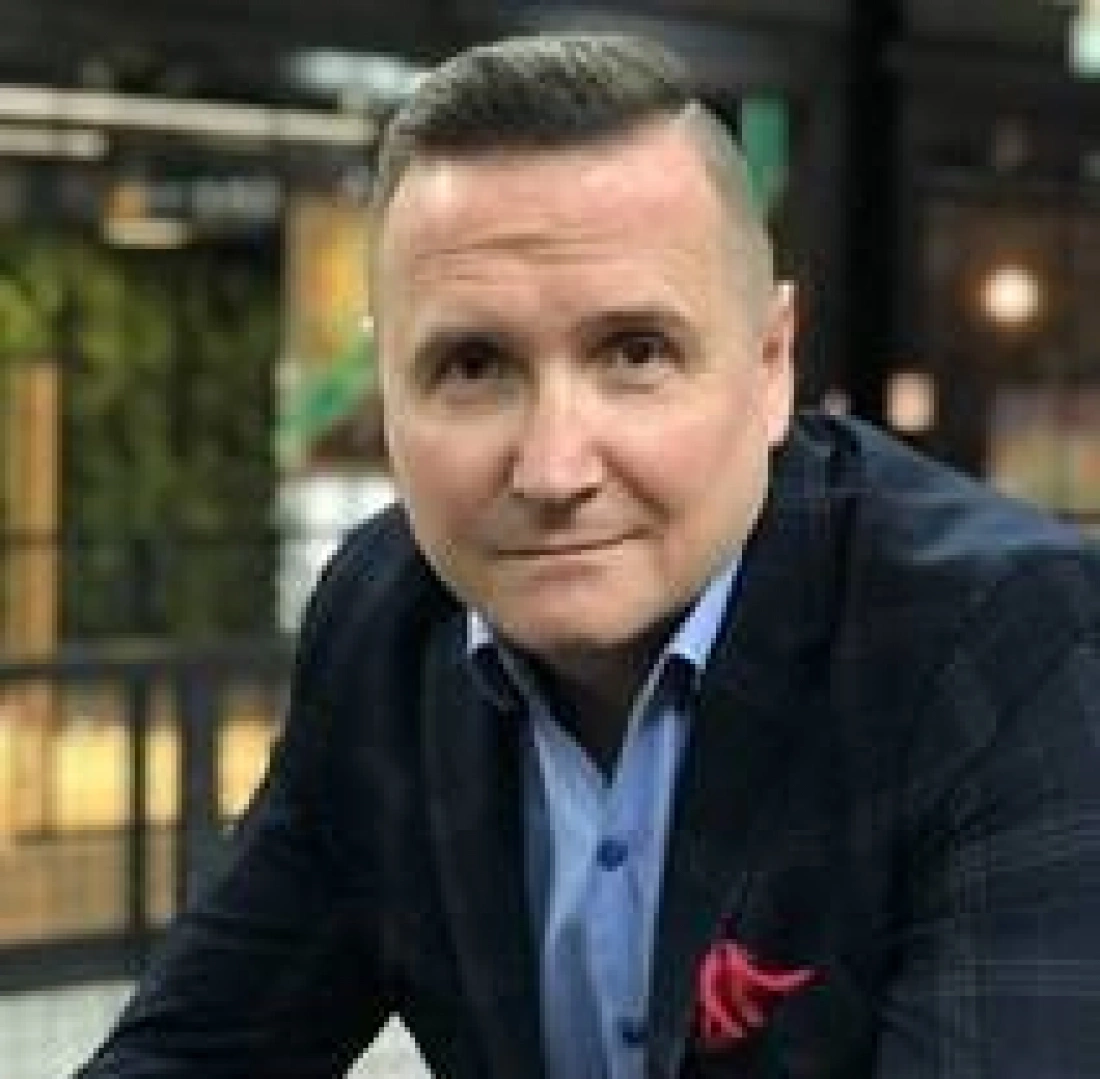 Mężczyzna w eleganckim płaszczu, z krótkimi włosami, uśmiechający się do kamery w nowoczesnym otoczeniu.
