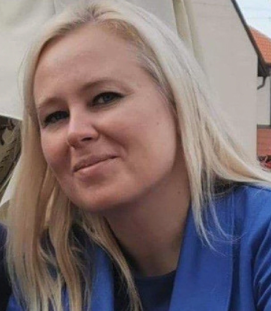 Kobieta z długimi blond włosami uśmiecha się, siedząc w przyjemnym otoczeniu.