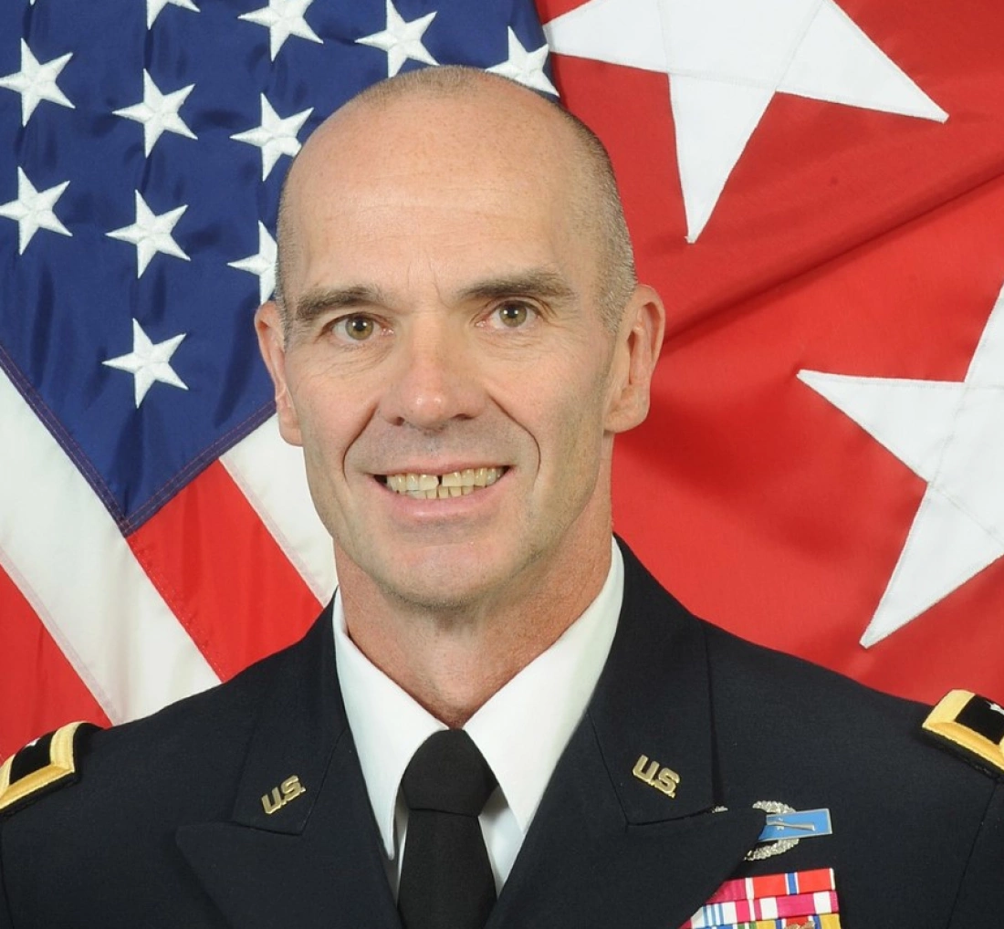 Oficer wojskowy w mundurze, z amerykańską flagą w tle.