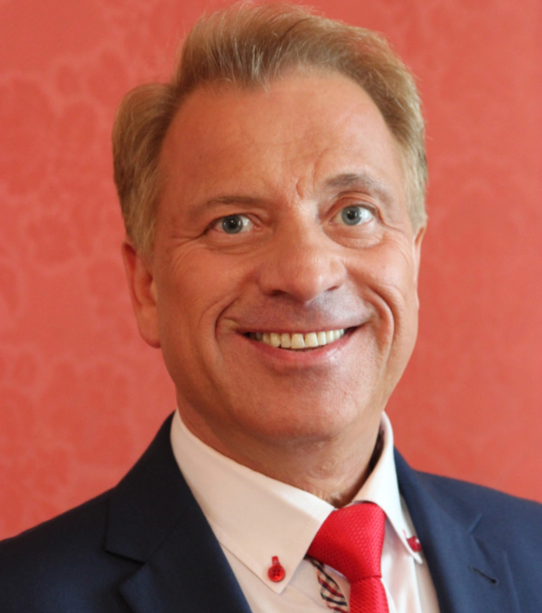 Mężczyzna uśmiechający się w eleganckim garniturze na tle o delikatnym wzorze.