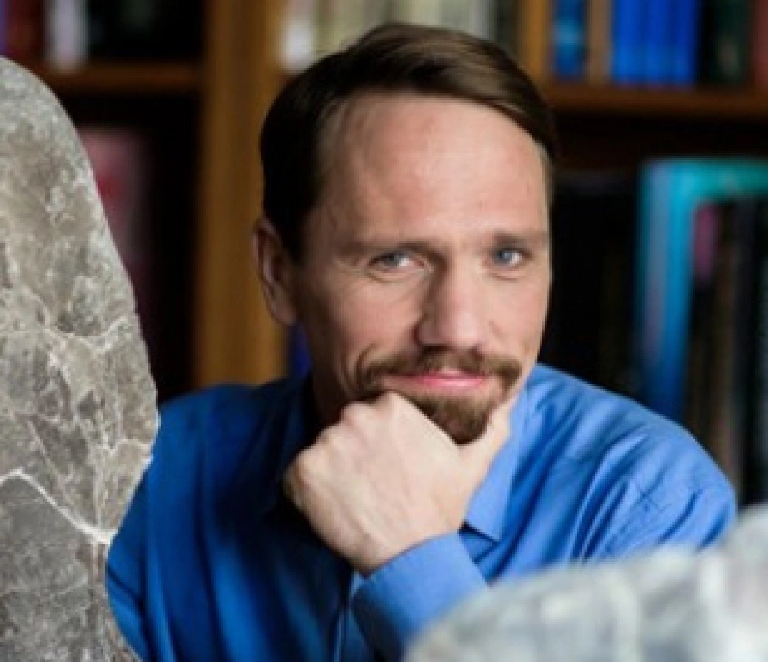 Mężczyzna w niebieskiej koszuli siedzi obok dwóch kamieni, z zamyślonym wyrazem twarzy. W tle widoczne regały z książkami.